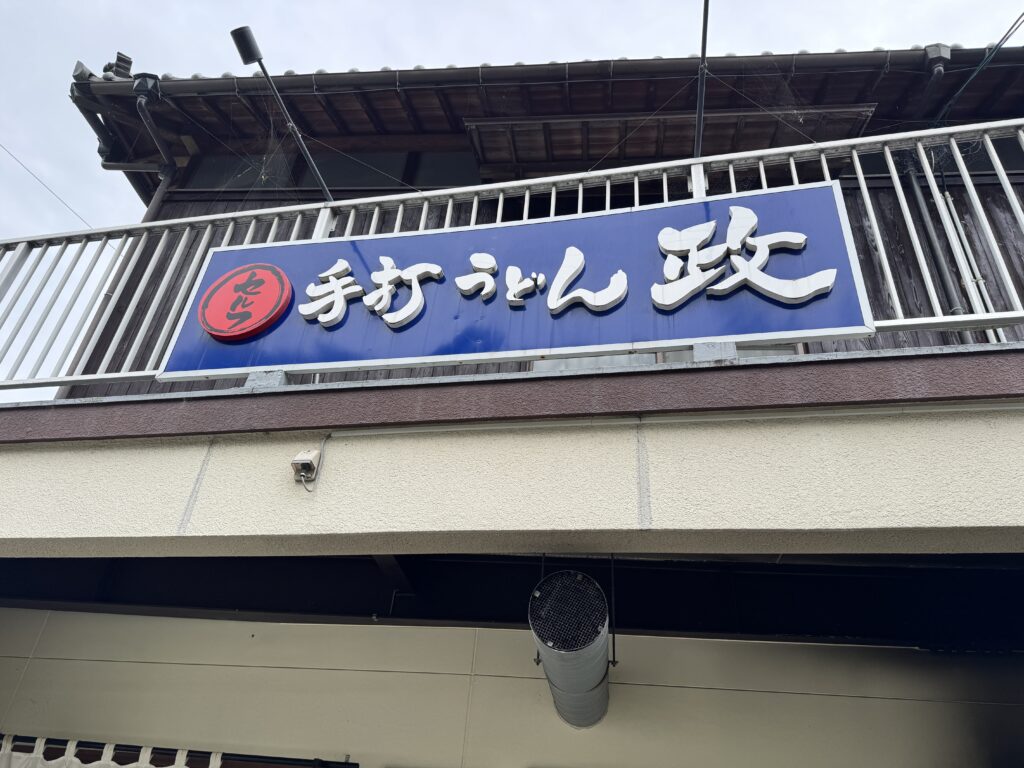 松山市の手打ちうどん店「政」の看板。セルフ表記がある青いサイン。